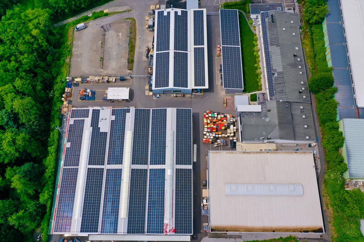 Luftaufnahme einer Industrieanlage mit Solarpanelen auf den Dächern, umgeben von grünen Bäumen.